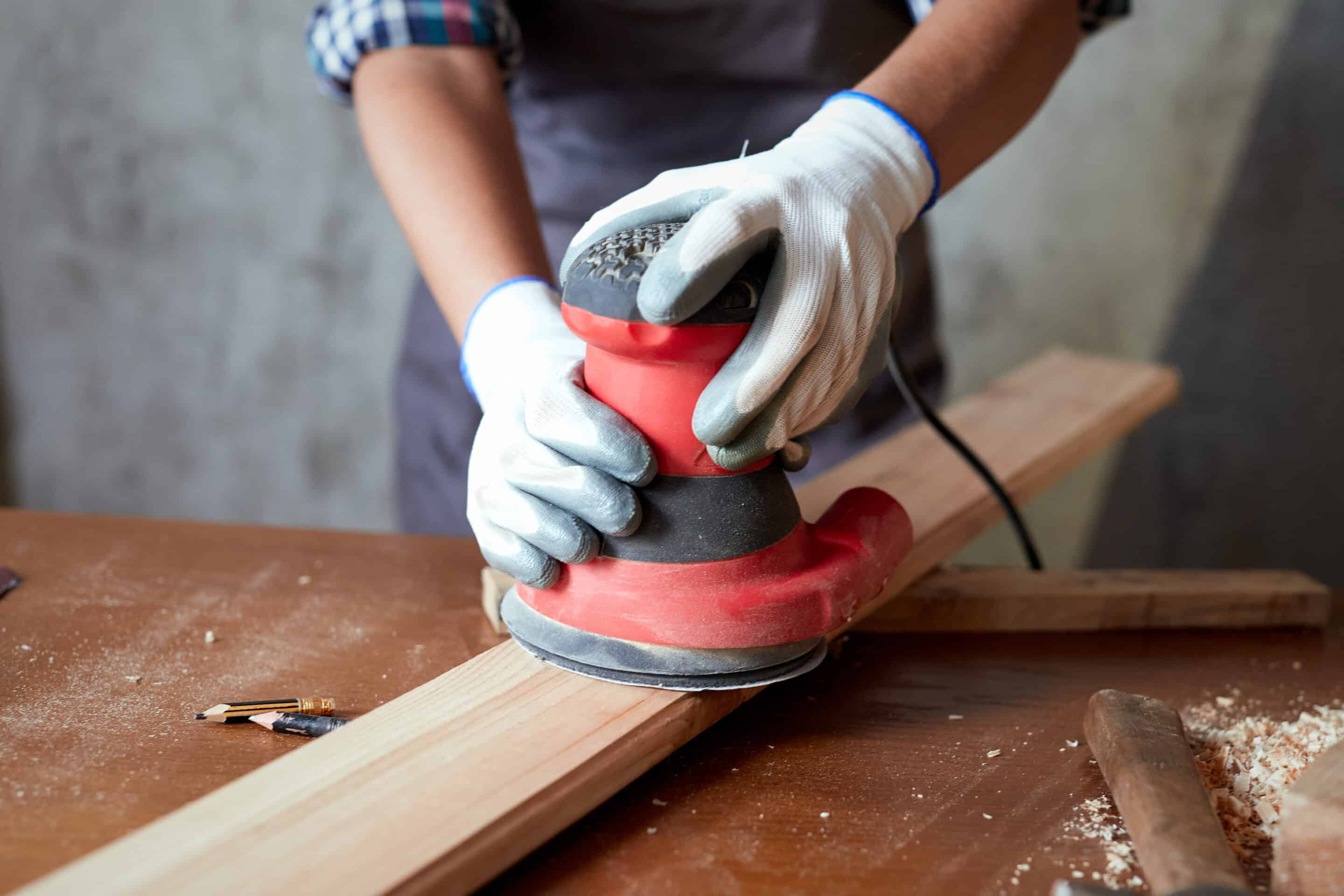 Female carpenter grinding wood with sandpaper in carpentry or diy workshop. Electric sander working in carpentry. Girl polishes wooden board with electric orbital sander.